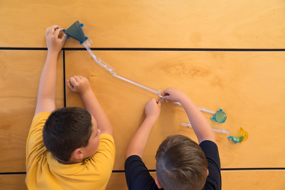 Two boys bent over, one in a yellow top, the other in blue, putting together a path out of plastic tubing.