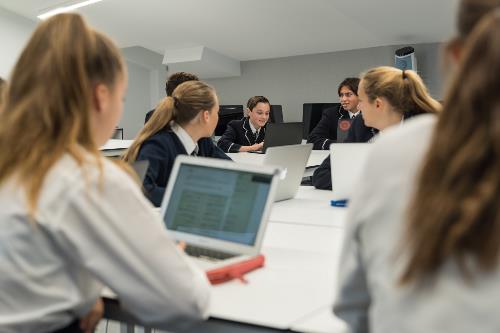Group of students, four blonde females in the foreground and three dark-haired students int eh background, collaborating with laptops open