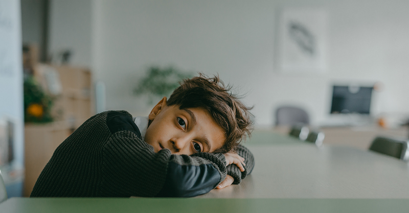 Young boy sits at desk with his head and arms resting on the table.