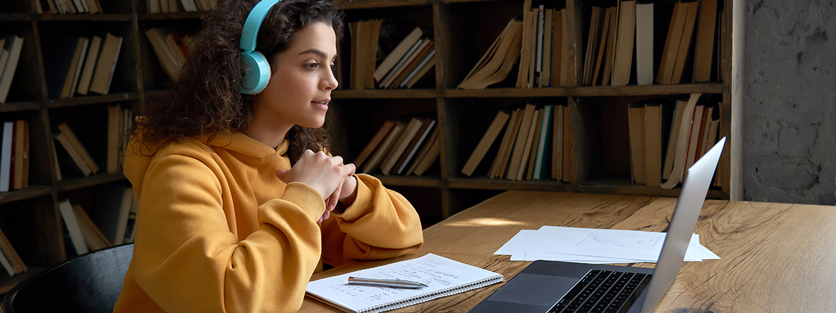 Teen Sits next to bookshelf, learning on a laptop.