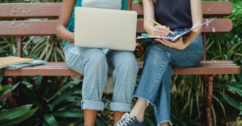 Two ladies sit on a park bench, one with a laptop and the other with a book. You can't see their faces.