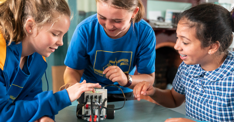 Three girls in uniforms sit around a table working on a robot.