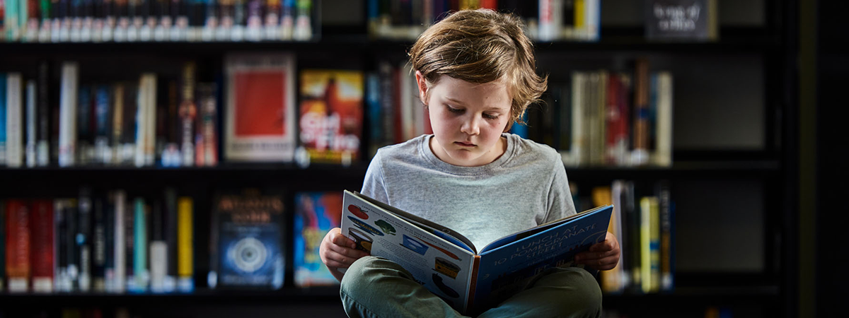 Child sits in front of library shelves, reading a book