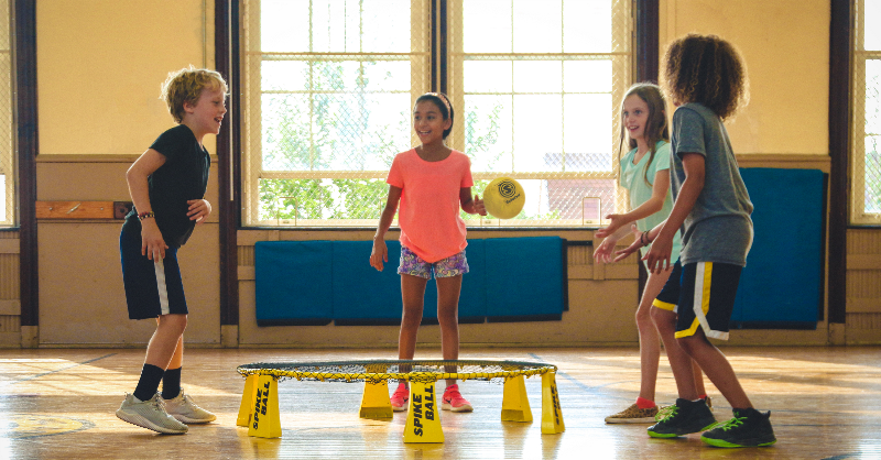 Four children standing in a circle with a trampoline between them, playing a ball game.