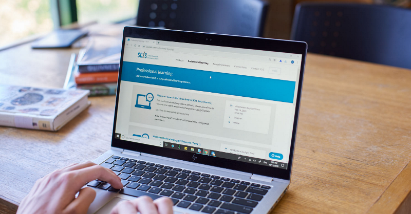 Someone working at a laptop on a desk, they're on the SCIS website. There's a coffee cup to the right of their computer and their mobile phone to the left.