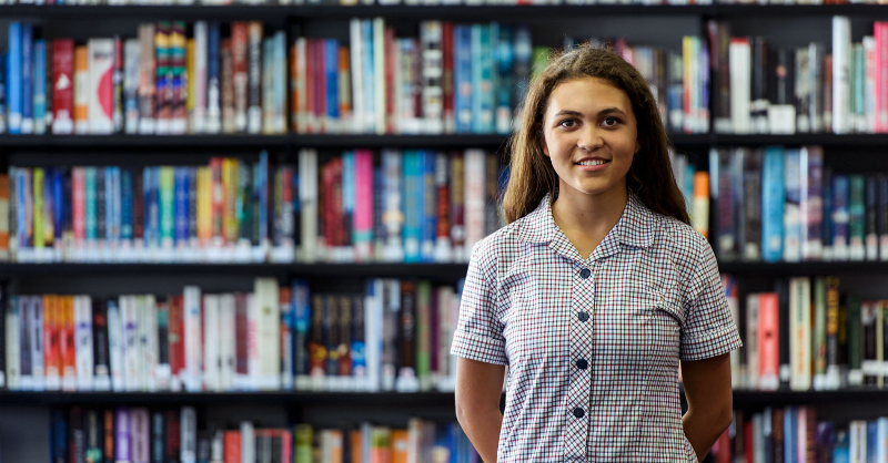 Young girl in school uniform, smiling at the camera, standing in front of shelves full of books. 