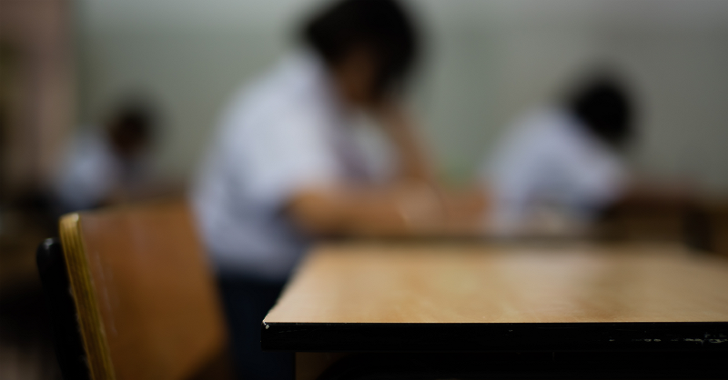 A wooden chair and desk sit empty in the foreground. Students are blurred in the background at their desks.