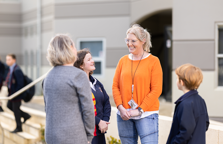 A teacher smiles and talks with a parent and two students outside a school building.